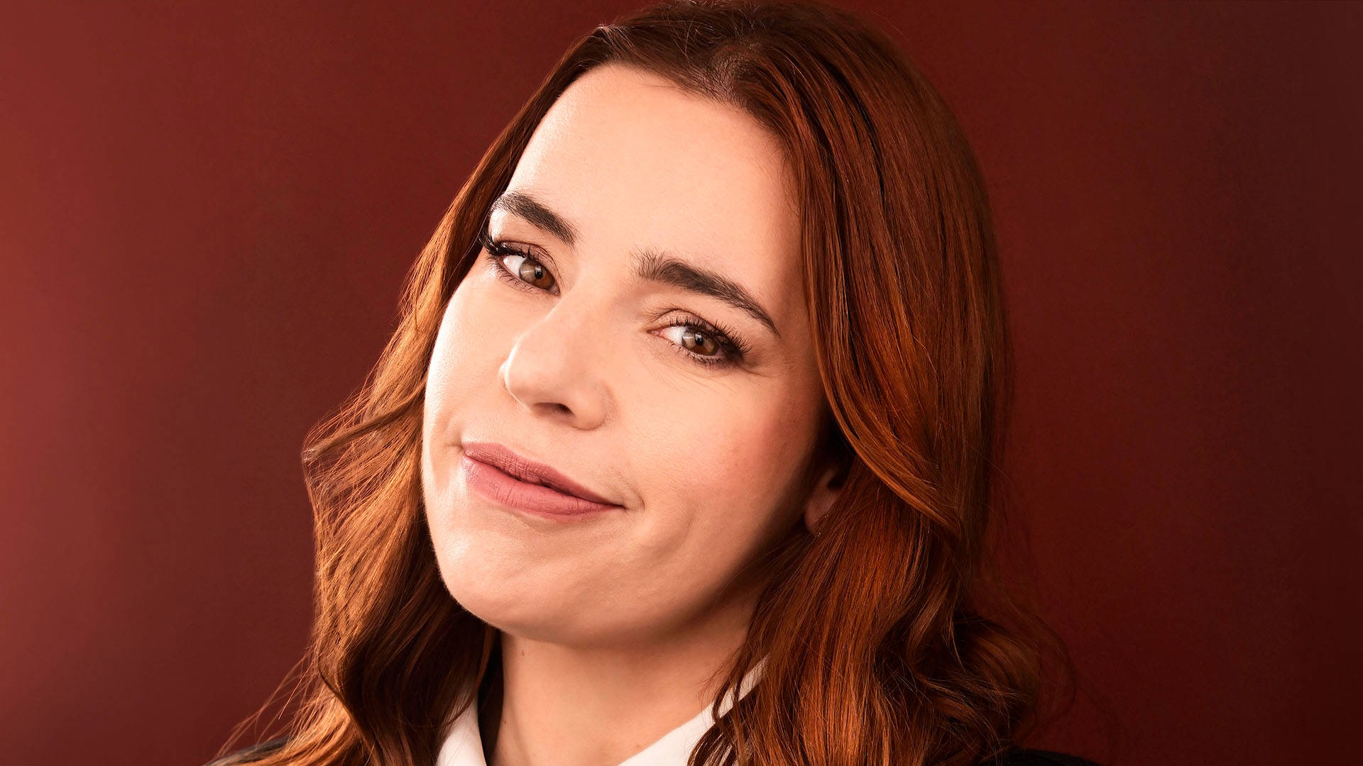 Alysha Umphress headshot: A caucasian woman with reddish-brown long wavy hair and brown eyes soft smiling in front of a deep red backdrop.