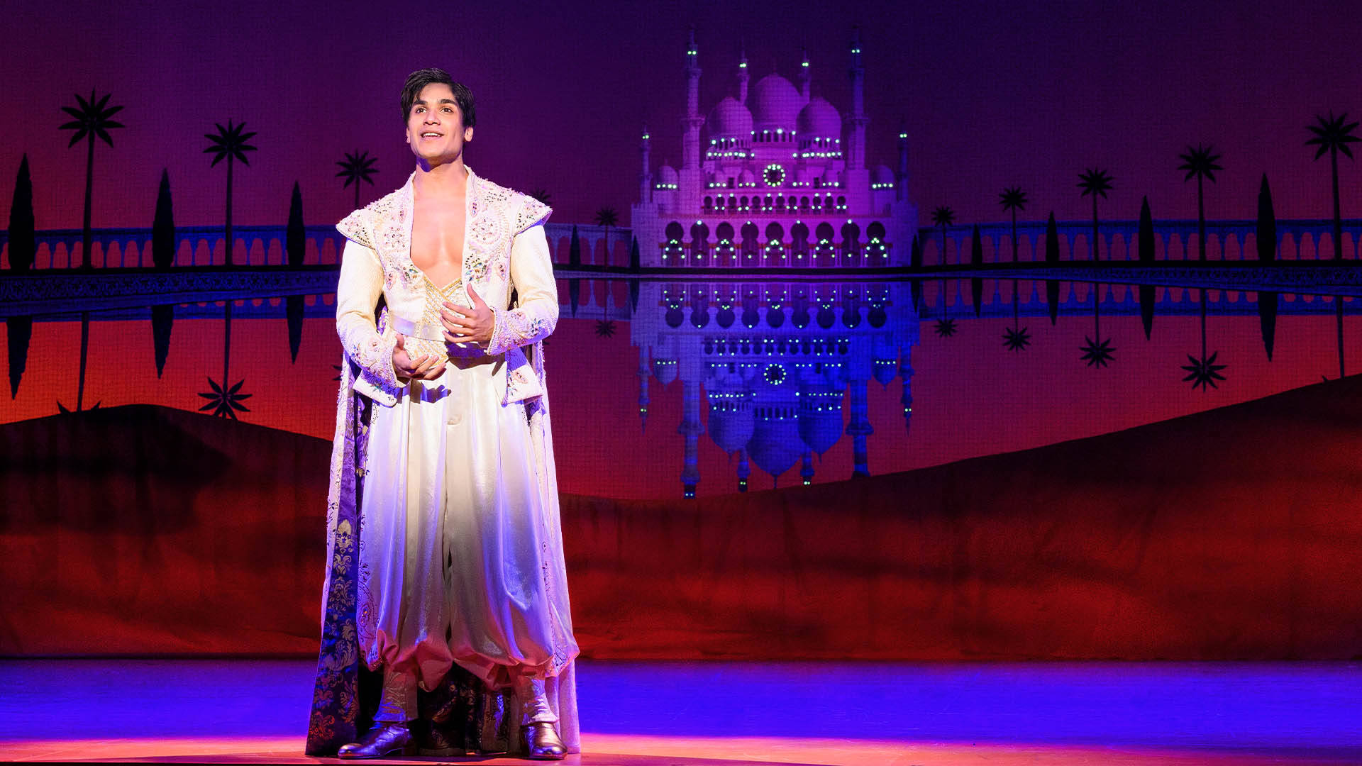 A man in a white, gem encrusted robe. He is staring out. Behind him is a giant palace on sand dunes.