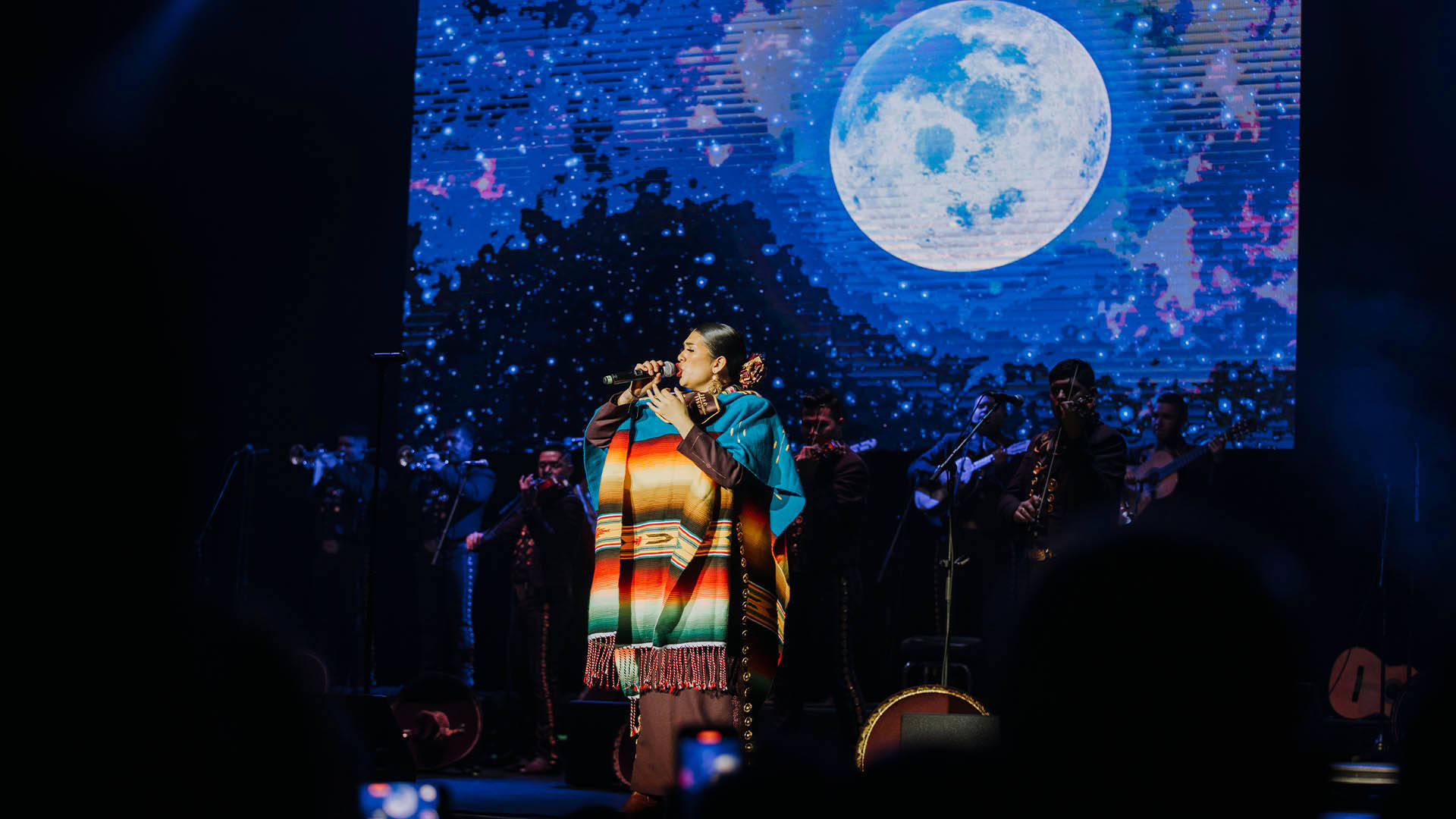 A singer in a bright, multi-colored poncho. She has dark hair and is holding a microphone. Behind her are other Mariachi band members, as well as a screen projecting a night sky with a large, full moon.