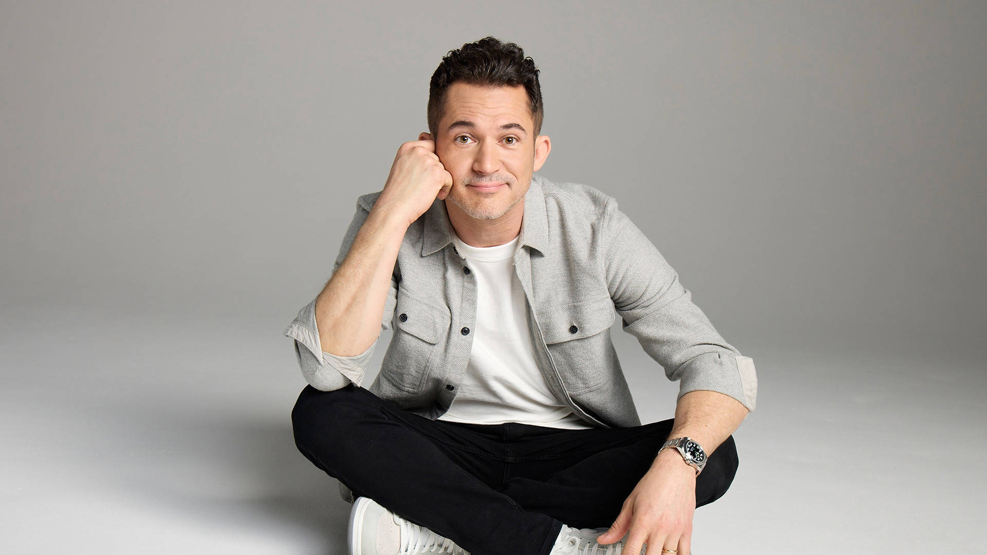 Justin Willman, a man with light complexion, short dark hair, wearing a grey shirt and black pants. He is cross-legged with one hand on his cheek, smiling softly.