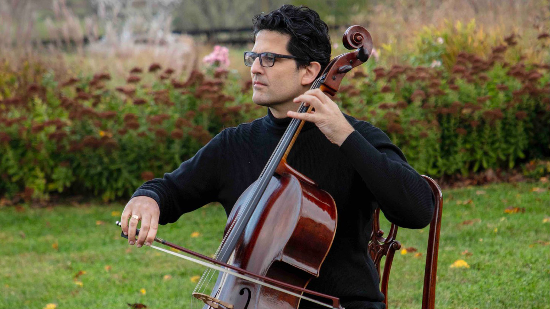 A cellist seated performing. He has medium complexion, short dark hair, and glasses.