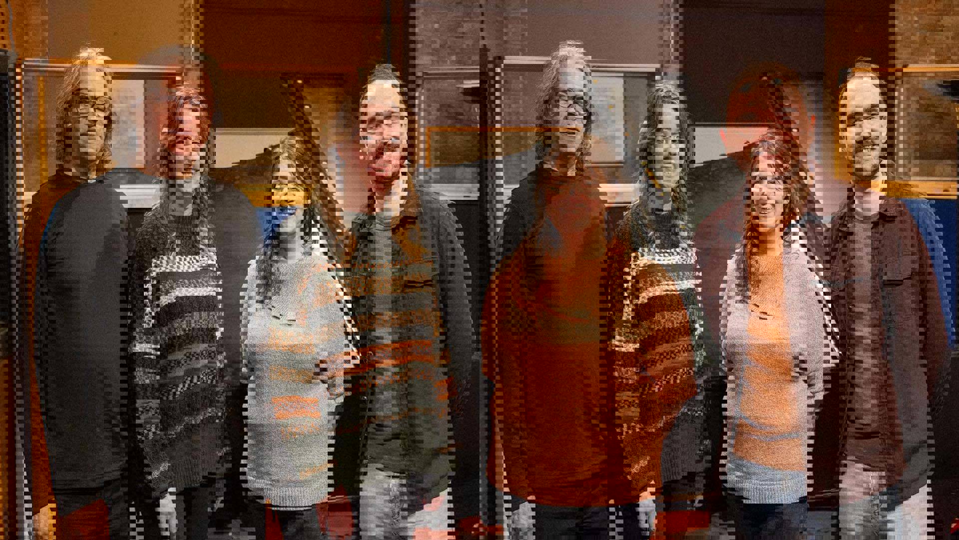 Four people stand together for a photo. The two men are on the outside and the two women on the inside. They all have light complexion and colorful clothing on. A piano is in the background.