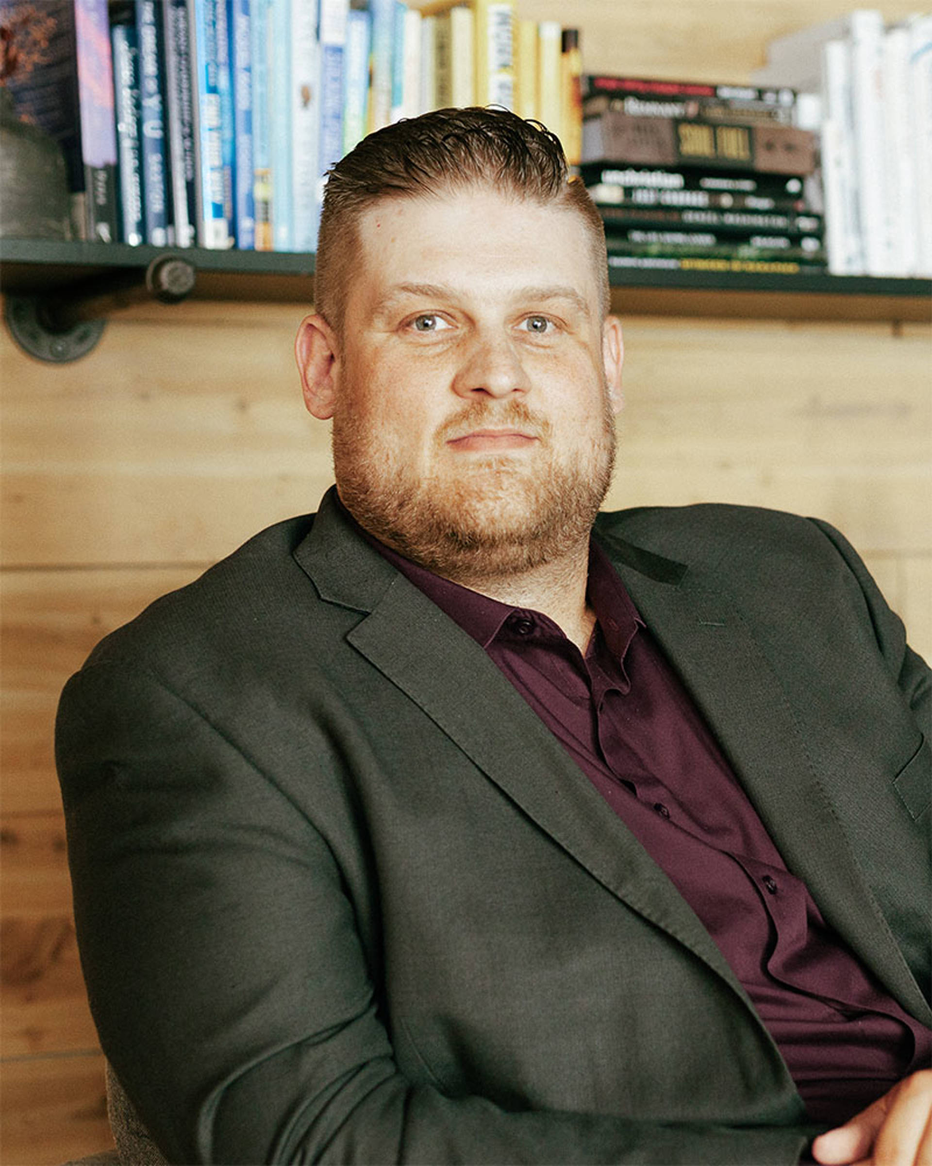 A young white man with reddish hair, a short beard and a grey suit jacket.