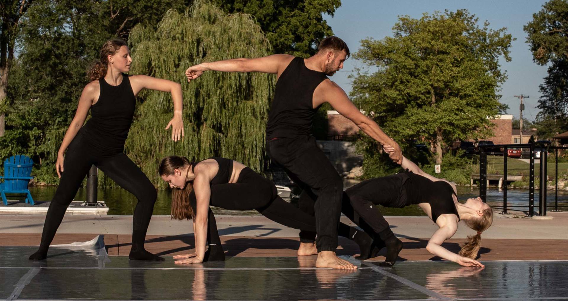 Four dancers in black on an outdoor stage. Three women are in various poses. One of them is being pulled to a standing position by a man.