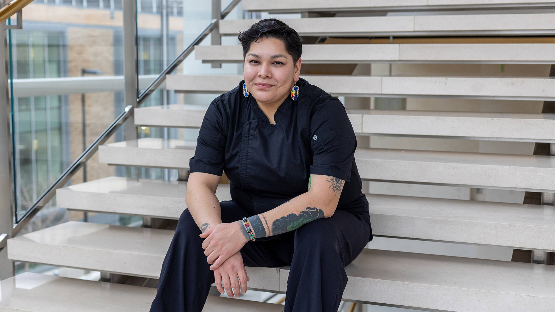 A medium complexion person with short black hair wearing a black chef coat and dangling blue and orange patterned circular earrings. She is sitting on stone steps and smiling, her arms showing a few tattoos, in Overture Hall Lobby.