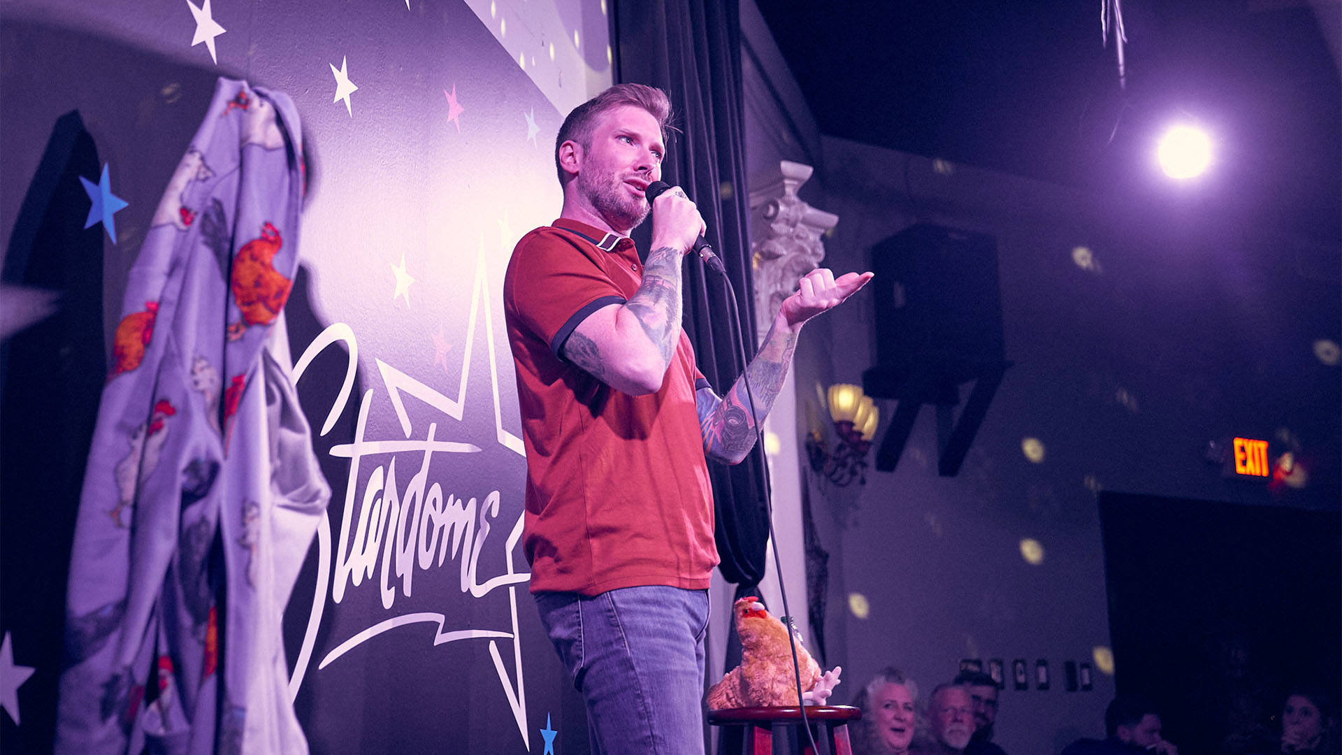 A young caucasian man with short blond hair in a red polo performing stand up on stage with a mic in hand.
