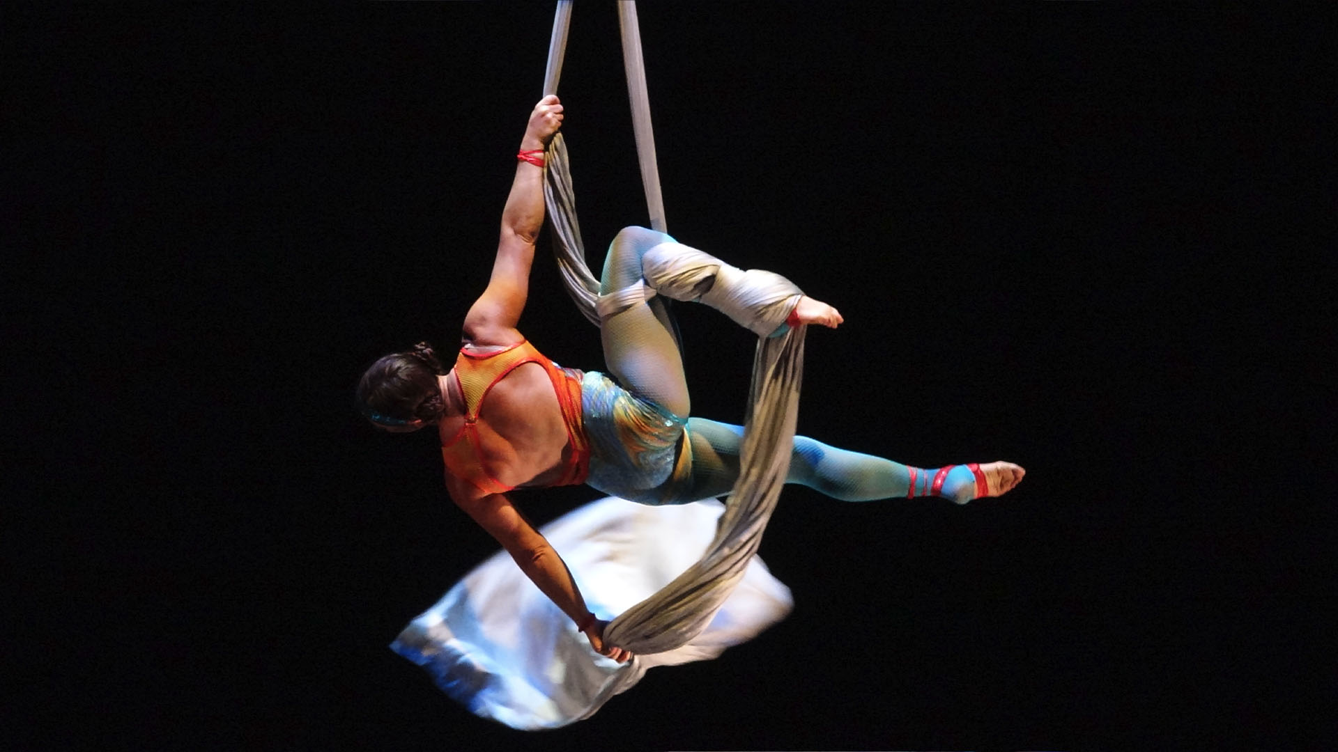 A trapeze artist hanging from a silver cloth. She is wearing an orange and blue leotard.