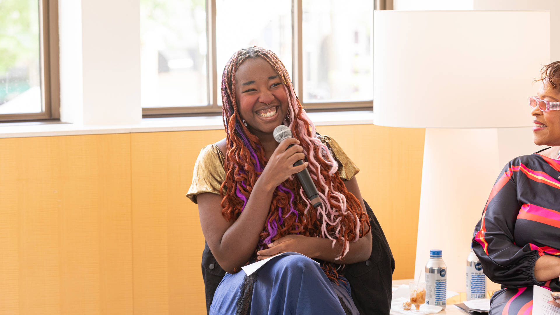 A young african american woman with long colorful hair sits and smiles while holding a microphone up near her face.