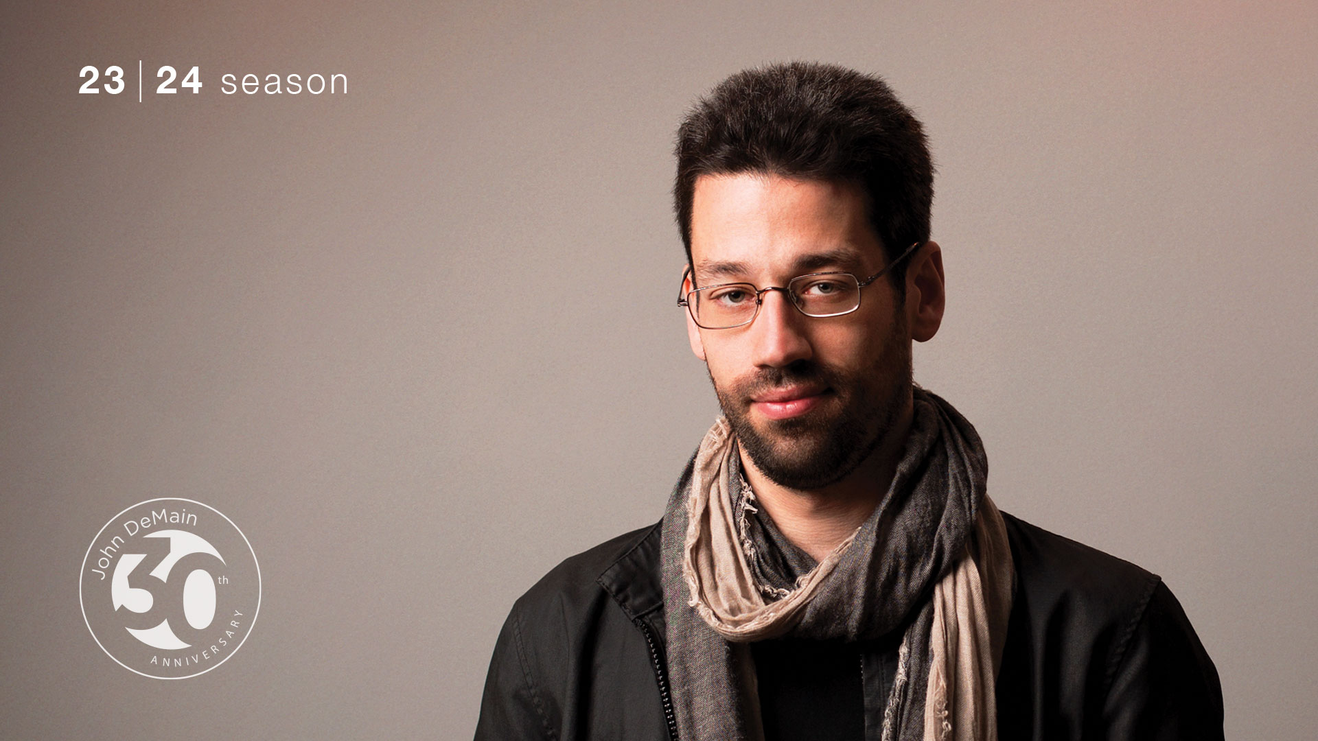 A young medium tone skinned man with short dark hair, a dark top, scarf and glasses against a tan background