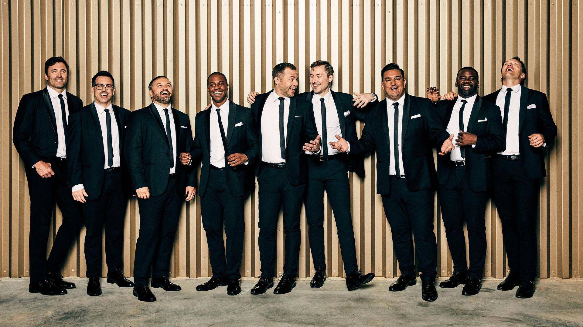 The nine members of Straight No Chaser stand in a line in black suits with black ties. They look like they're having a great time and are all smiling. A couple are laughing.