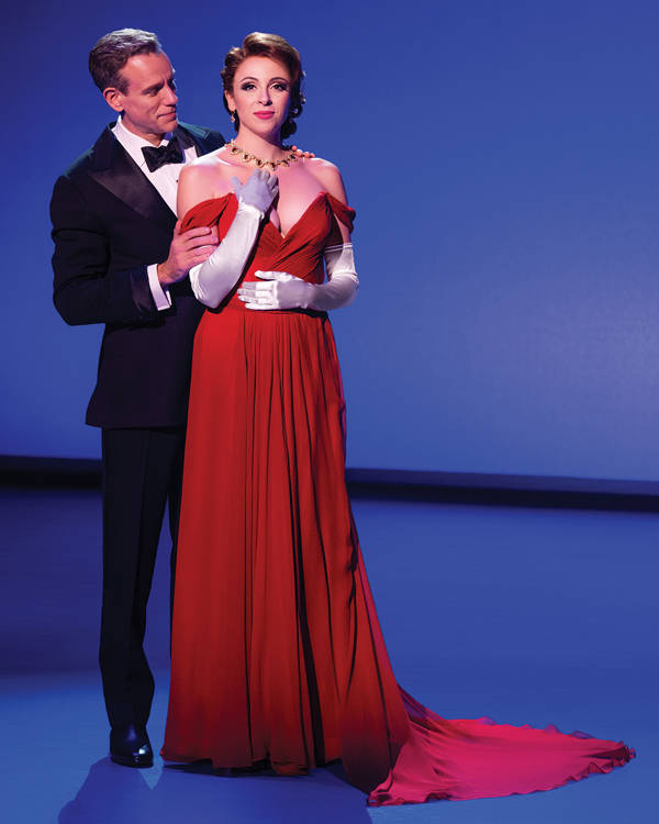 A man and woman with light skin stand together. He is wearing a dark suit and she is wearing a red dress.