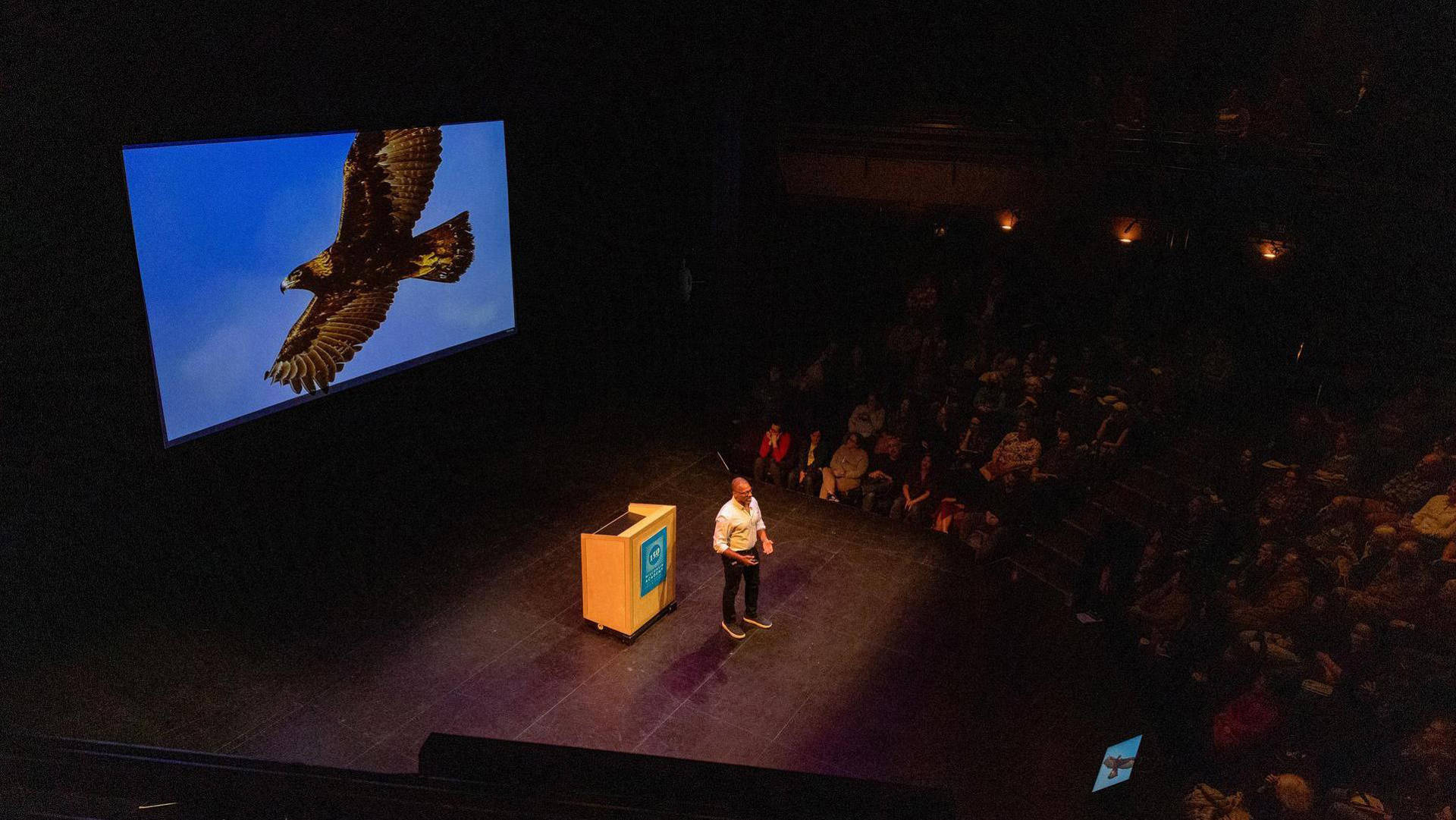 An african american man with buzzed greyish hair, glasses, and a white button-up shirt speaking to a seated audience. Behind him is a projection of a large bird against a blue sky.