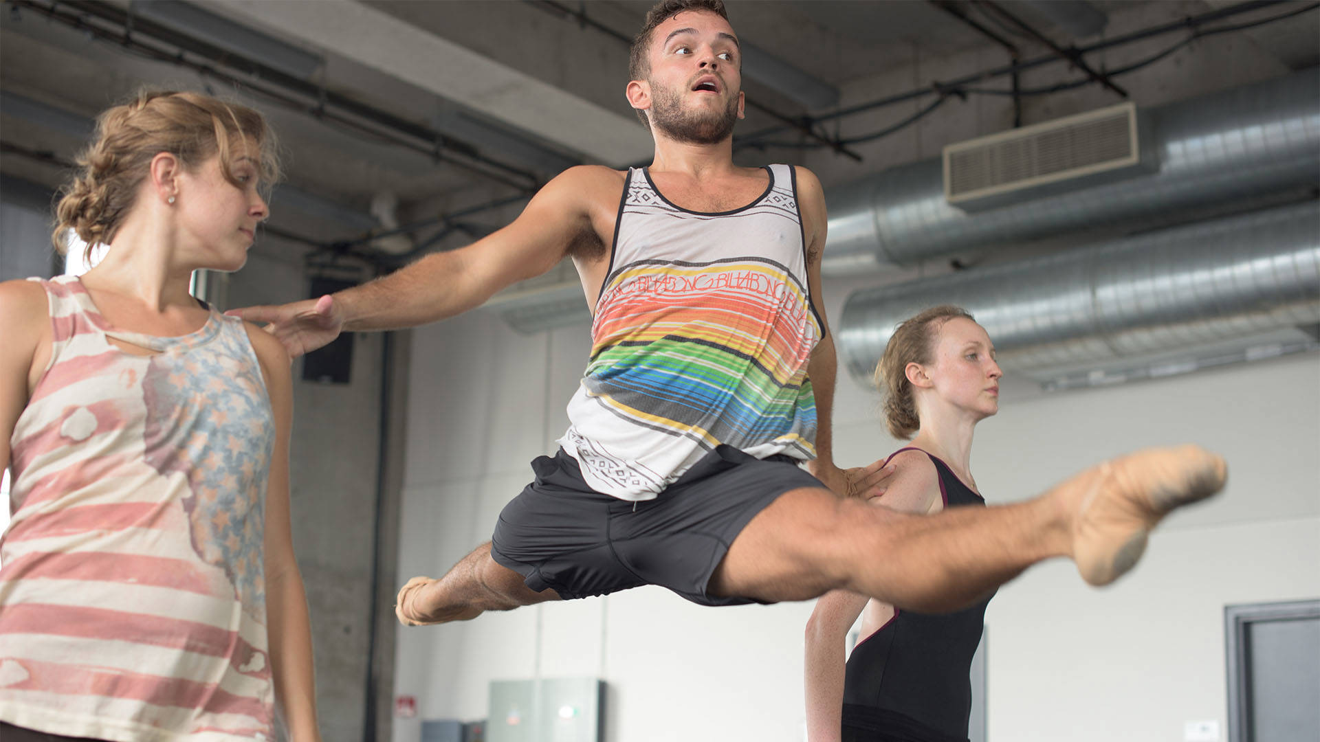 Three dancers in a studio. The main subject is a light complexion person in a tanktop and shorts with brown short hair and a light beard who is mid-leap with ballet flats on.