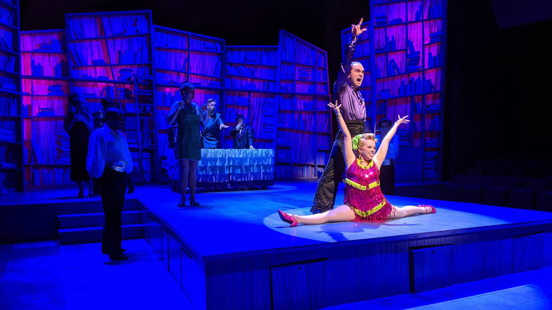 Performers on a dark theater set with a spotlight. In the center, a man and a woman gesture towards the audience in glittery outfits. The woman is doing the splits.