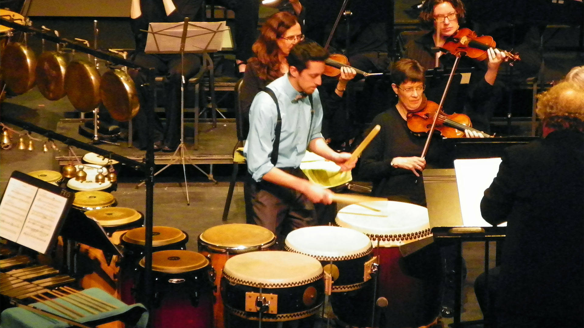 A young caucasian man with short dark hair, and blue dressshirt, and suspenders playing a multi percussion setup in front of three violin players in dark outfits
