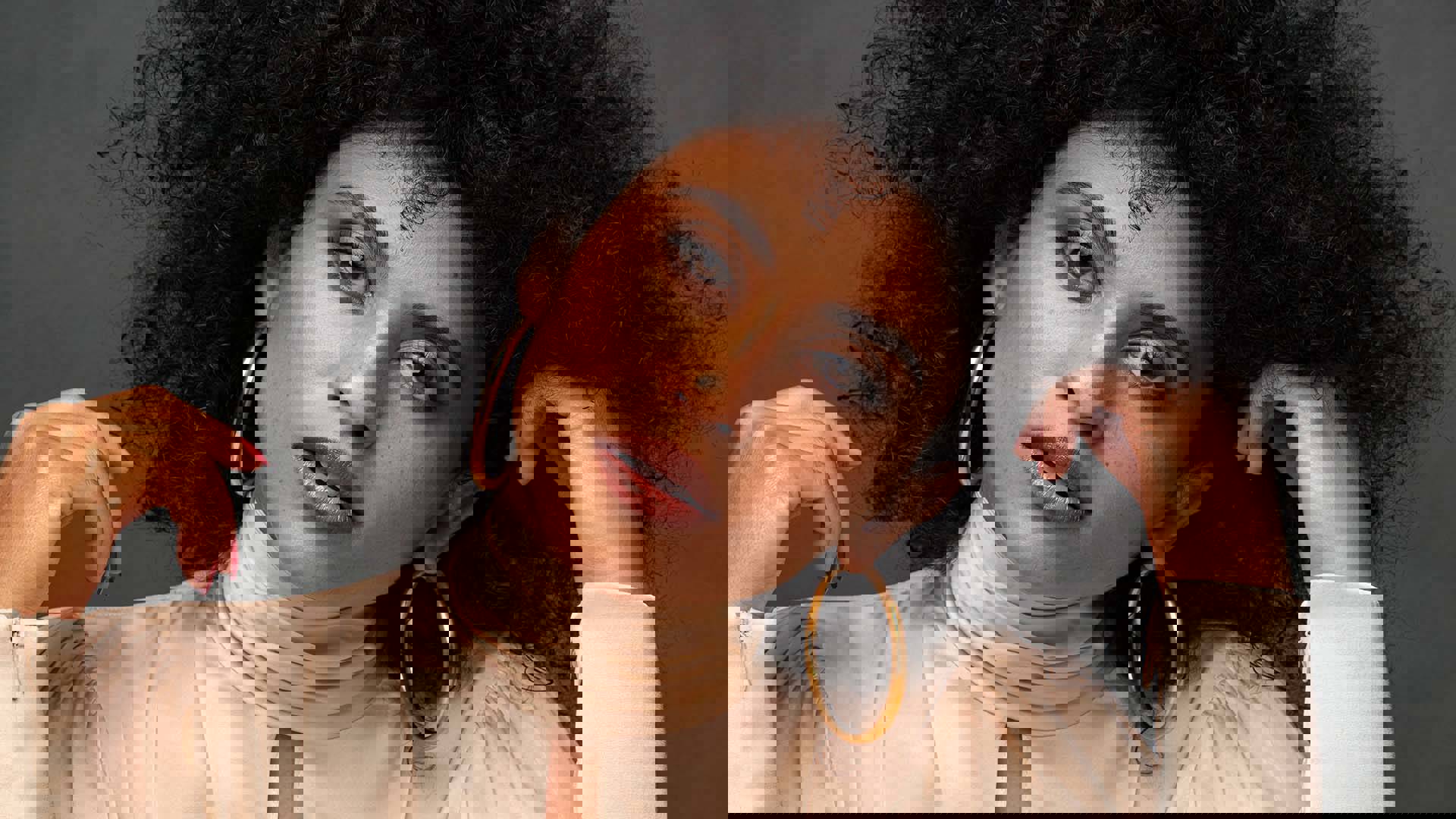 Alea looks at the camera and leans her head to her left. She has brown eyes, dark curly hair and gold earings on. One hand is up playing with her hair.