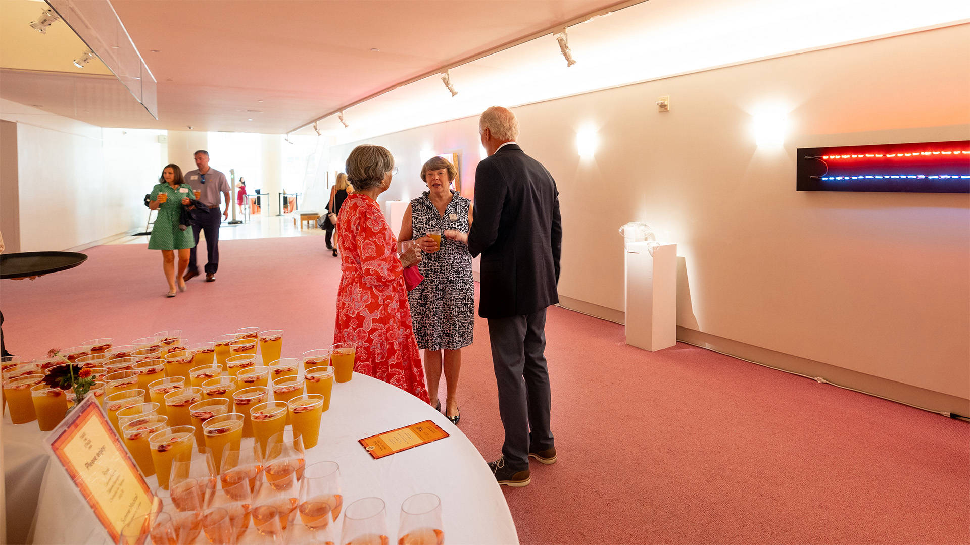 A woman with light complexion in a white and black dress talks to two others in an art gallery with pink carpeting.