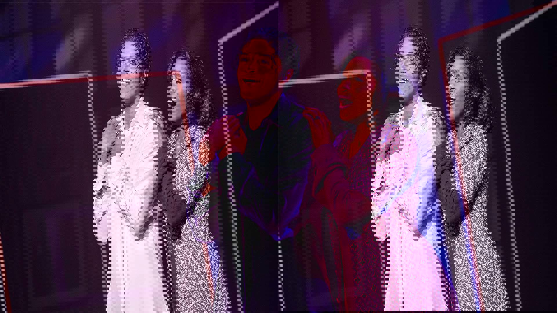 An actor playing Michael Jackson smiling and looking towards the left side of the frame. He has medium complexion and dark hair. A woman stands beside him with her hand on his shoulder. She has dark complexion and dark hair.