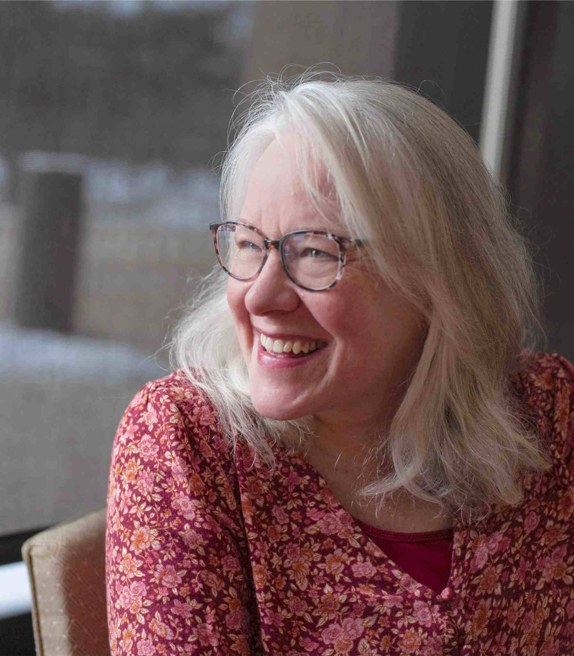 Headshot of a smiling middle-aged white woman with grey hair, glasses, and light blue eyes. She is wearing a floral red top.