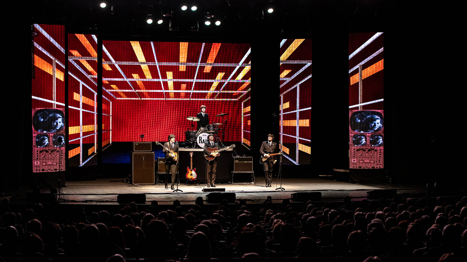 Four Beatles impersonators performing in front of a full house. They are wearing dark suits with ties and holding guitars.