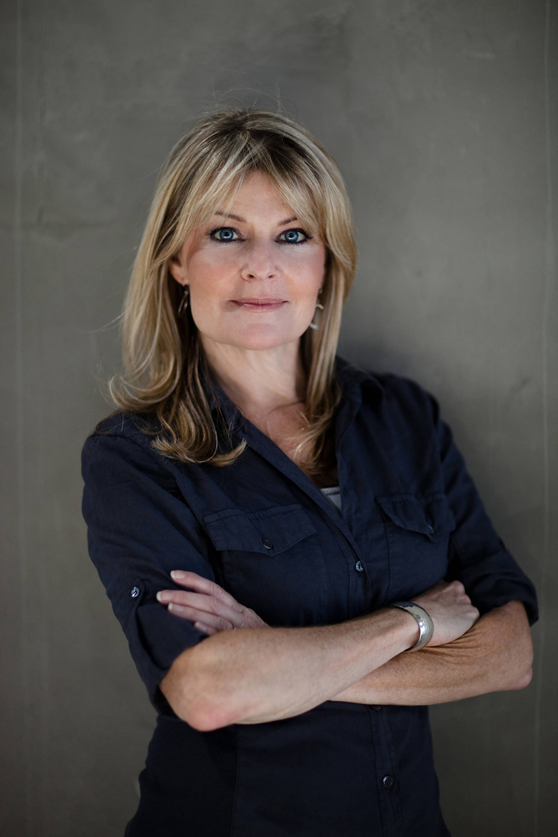 Deanne Fitzmaurice. A caucasian woman with long blonde hair and blue eyes. She is wearing a navy blue button up shirt rolled up to her elbows with her arms crossed against a grey wall.