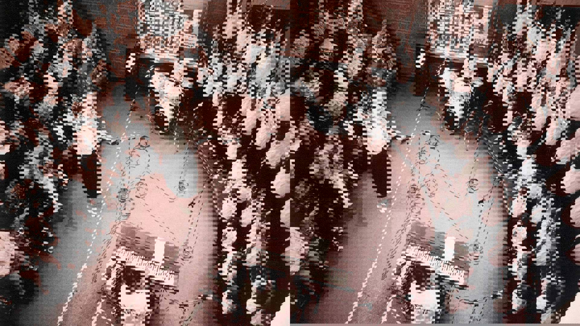 Greyscale photo of a childrens choir with their arms raised, a director, a drum set player and keyboard player performing for a seated audience. The shot is from above.