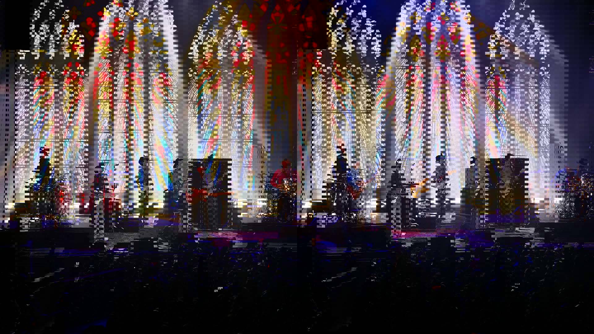 Intocable, a six-piece band performing on stage with stained glass windows and lighting behind them.