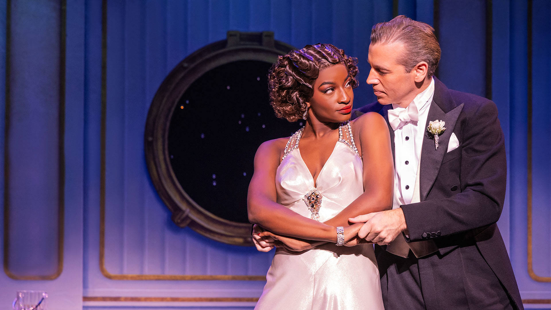 A black woman with brown, curled chin length hair pinned up wearing a silky white dress with pearl accents. She stands next to a light complexion man with greying hair in a tuxedo. They are looking into each other's eyes mid-twirl while dancing.