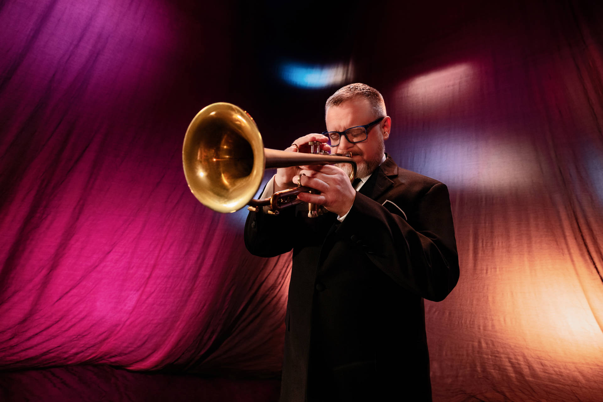 A man with light complexion and light brown hair plays a trumpet in front of a pink stage curtain.