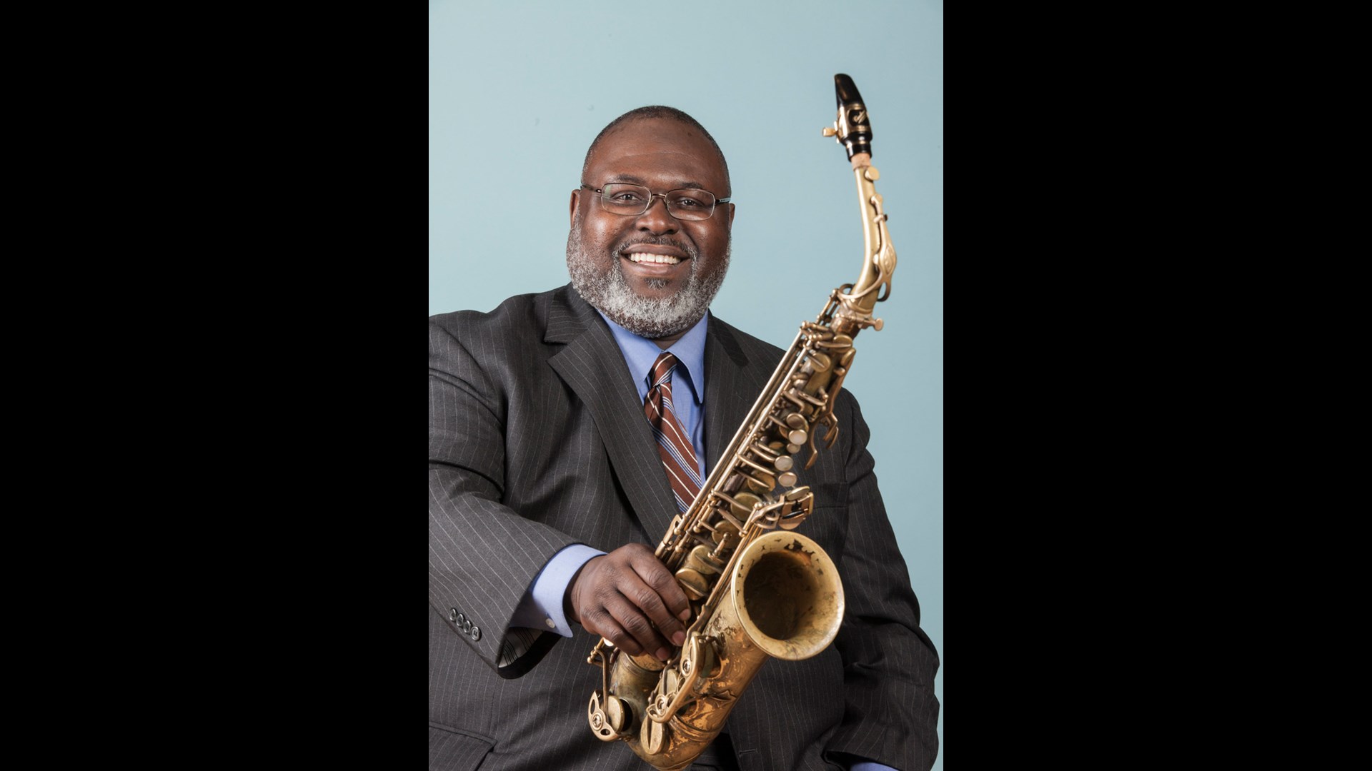 A man with dark complexion, a grey beard and glasses smiling to the viewer and holding out an alto saxophone. The background in blue.