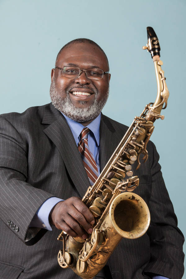 A man with dark complexion, a grey beard and glasses smiling to the viewer and holding out an alto saxophone. The background in blue.