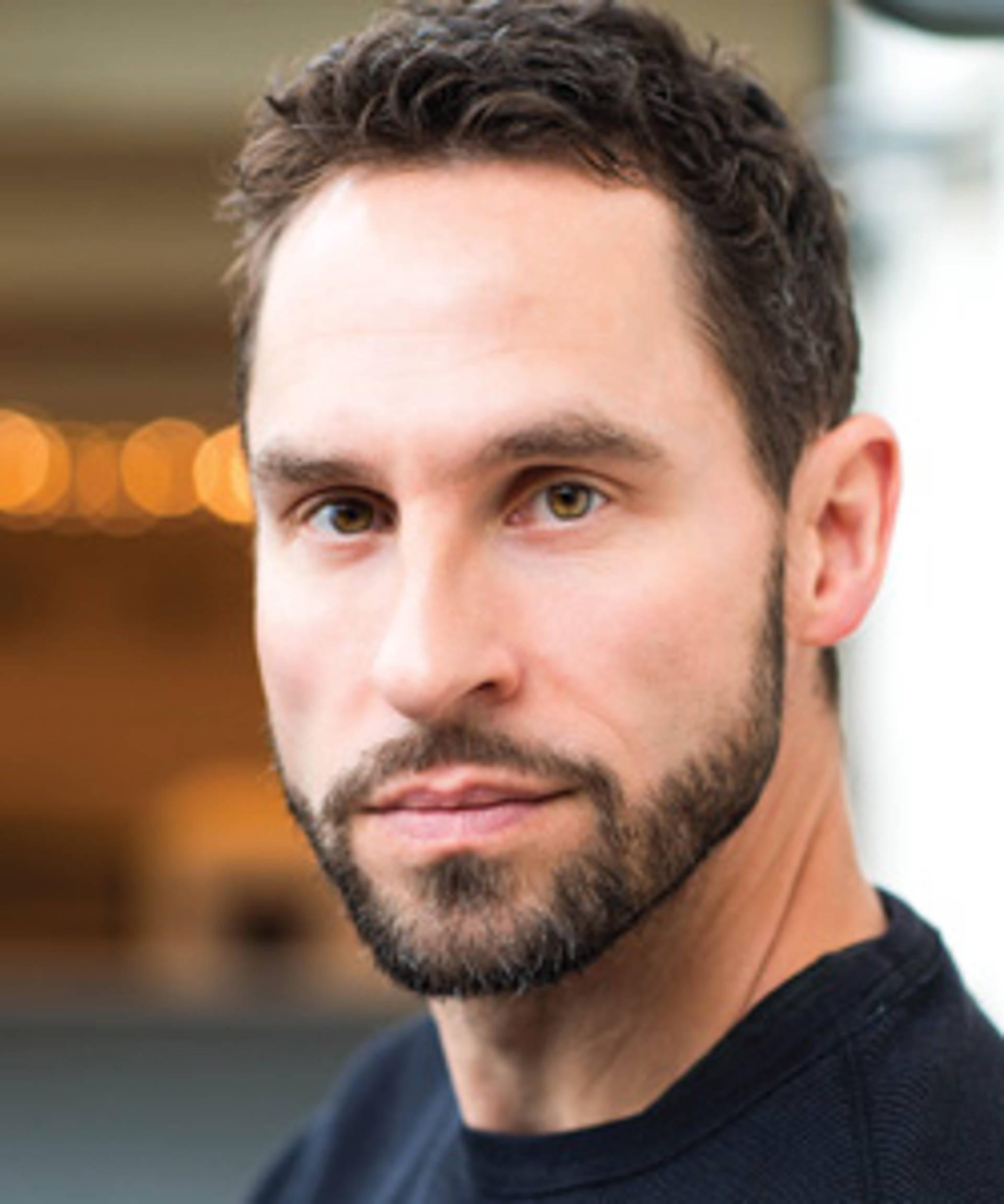 A light skinned man with a short dark beard, dark hair and dark eyes.