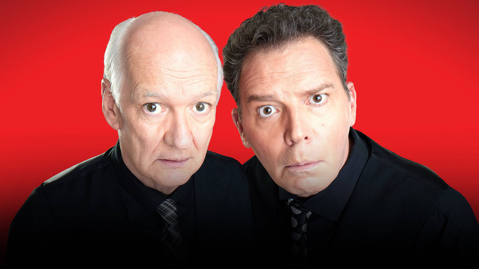 Colin Mochrie and Brad Sherwood, two older white men standing close together wearing black suits with dark ties. They are looking up into the camera with curious faces on display in front of a red background.