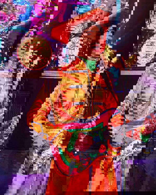 A George Harrison impersonator wearing an orange Seargent Peppers outfit and holding a guitar. He has brown hair and a moustache.