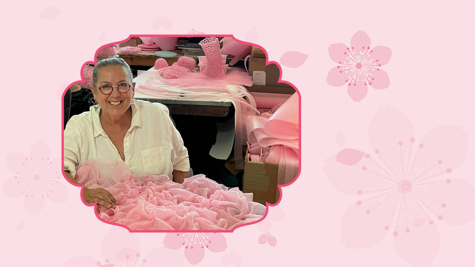 Pink Banner with a middle aged woman with light complexion, grey hair, glasses and a white blouse. Piles of pink fabric are all around her.