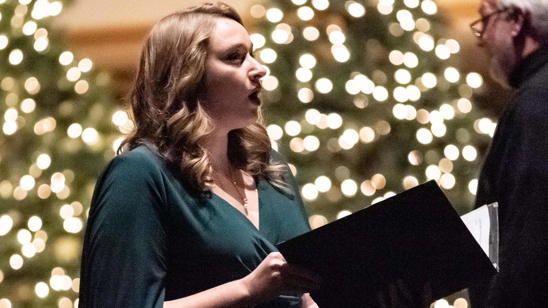 A white woman with blonde, shoulder-length hair in a green dress. She is singing and holding a choir book. Behind her are Christmas trees with lights.