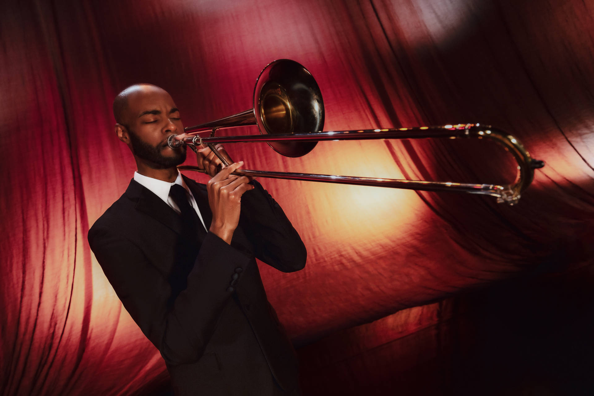 A man with dark complexion plays a trombone in front of an orange stage curtain.