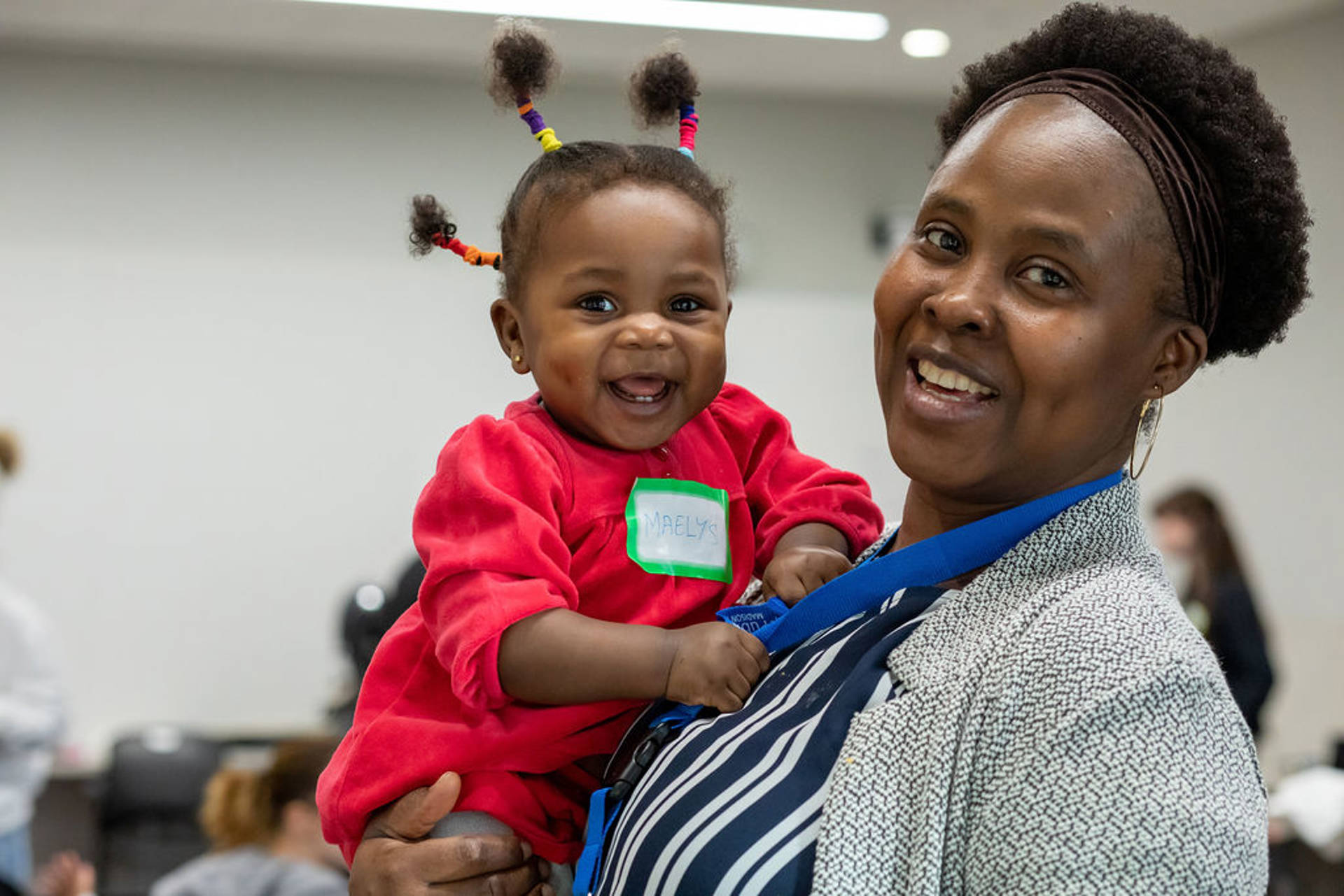 A woman with dark complexion and brown eyes holds a toddler. They are both smiling towards the viewer.