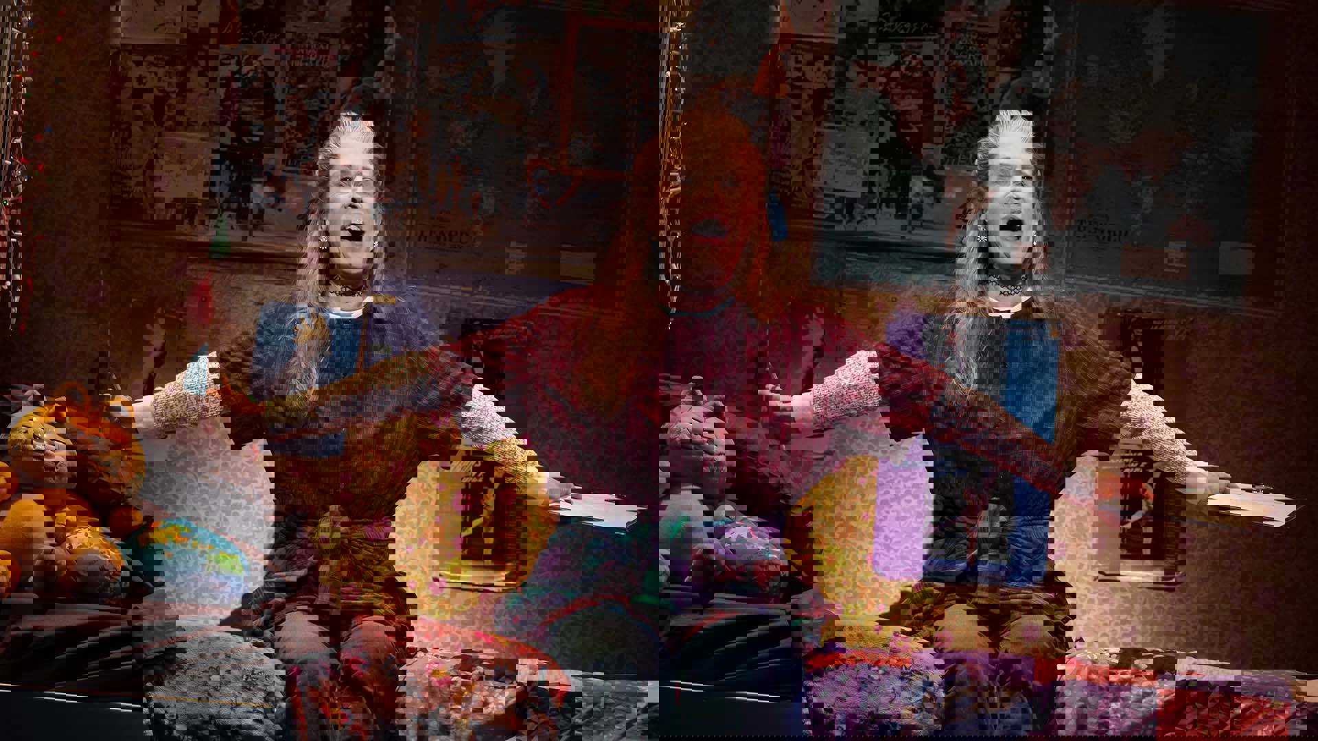 Woman with light complexion sits on bed and sings with arms outstretched.