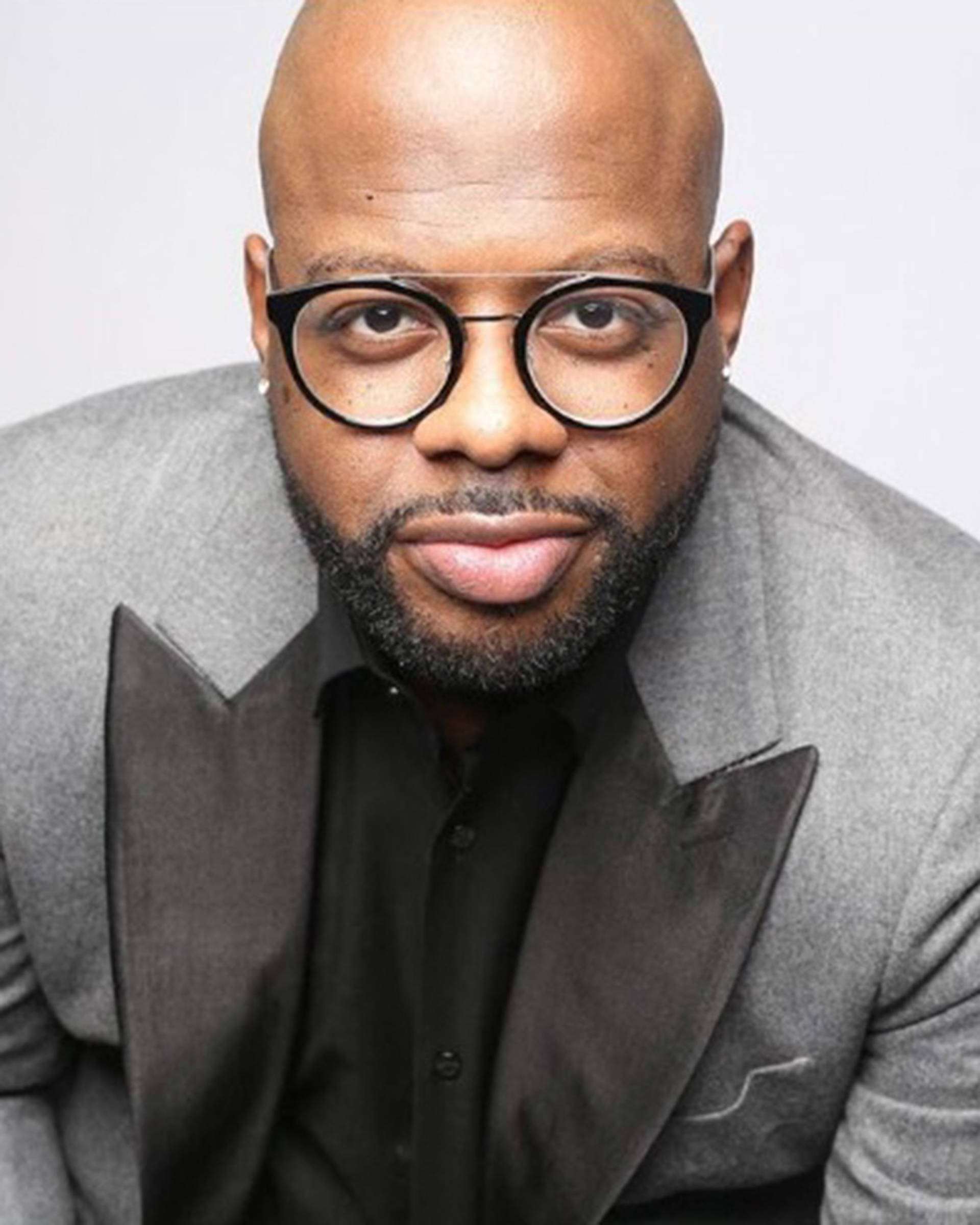 A headshot of an African American man in a silver and black suit with black glasses and a beard