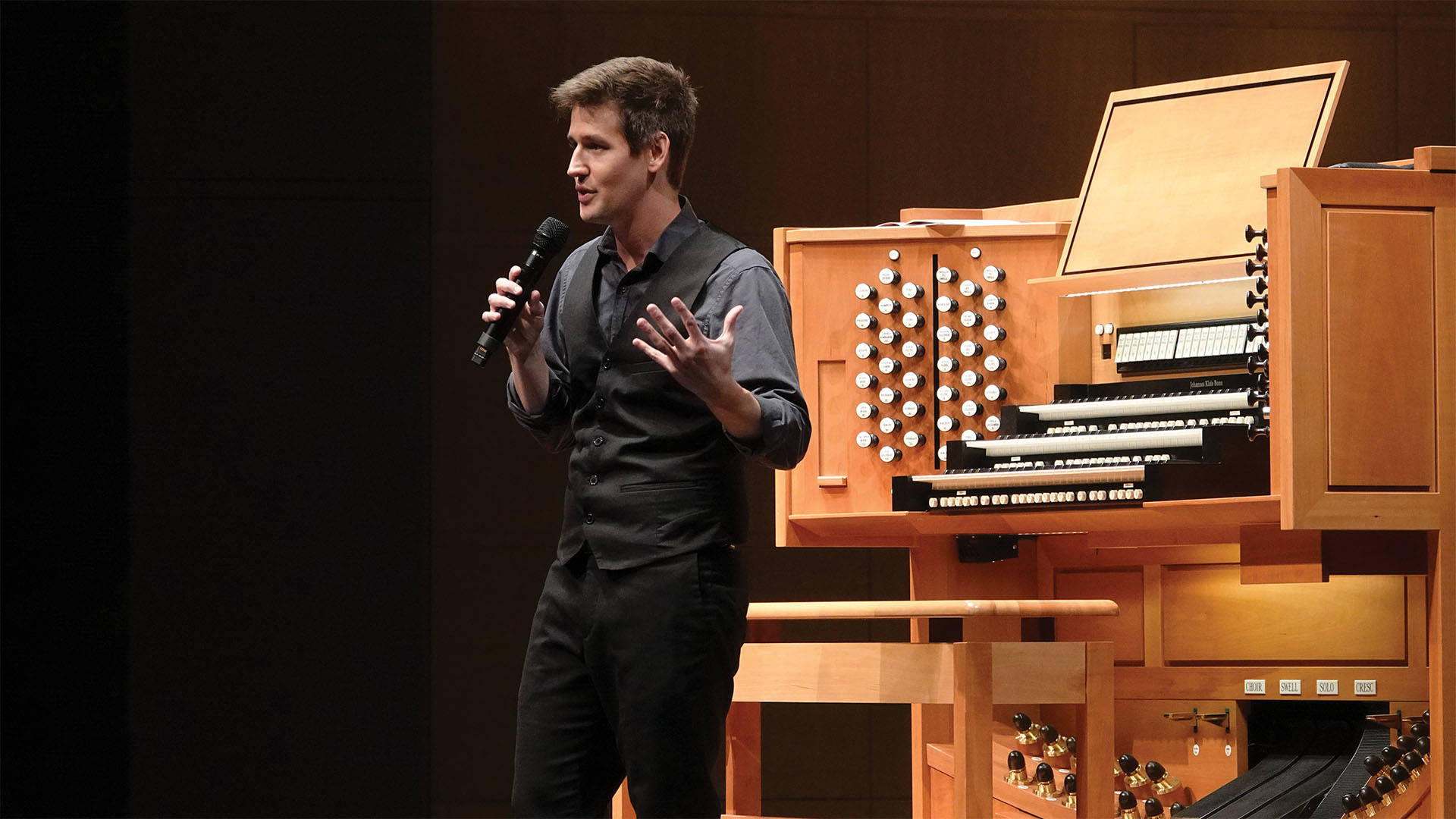 Greg Zelek, a young man with light complexion, short styled brown hair, a black button-up shirt and black vest, speaking into a microphone in front of a large wooden console.