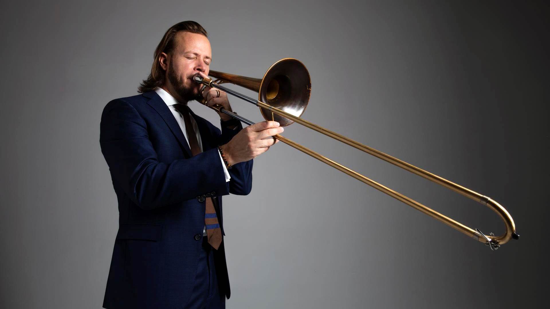 A white man with long blond hair and a blue suit playing trombone against a grey background.