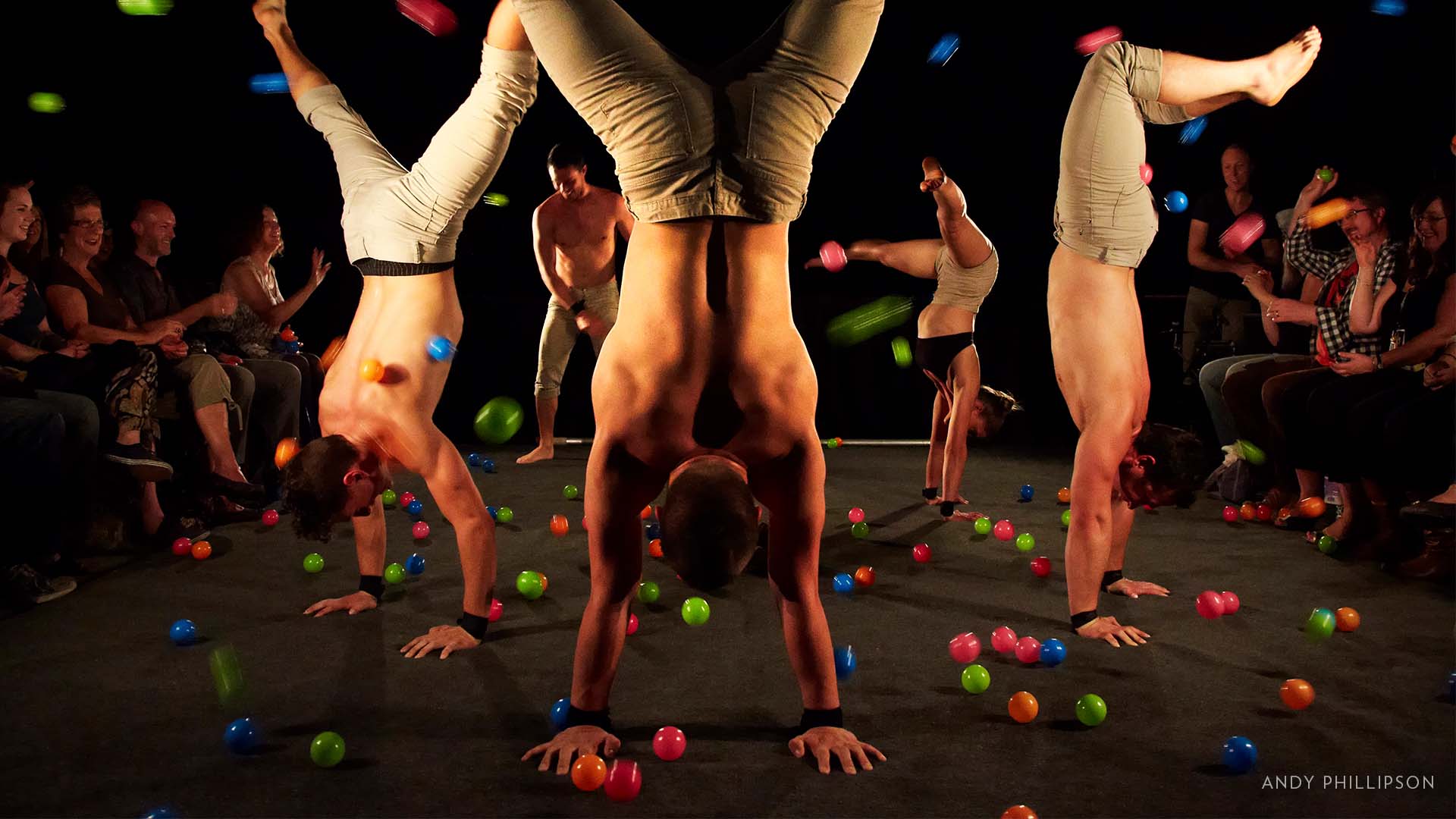 4 dancers with minimal upper body clothing and tan pants on are doing handstands in front of a close audiences. There are colorful balls everywhere, and flying through the air.