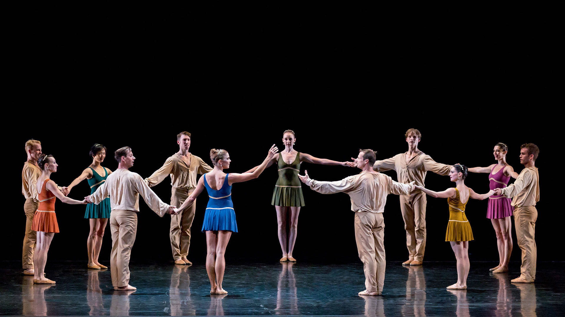 An ensemble of dancers in tan and earth tone outfits standing in a circle on stage with their arms out on both sides to hold hands.