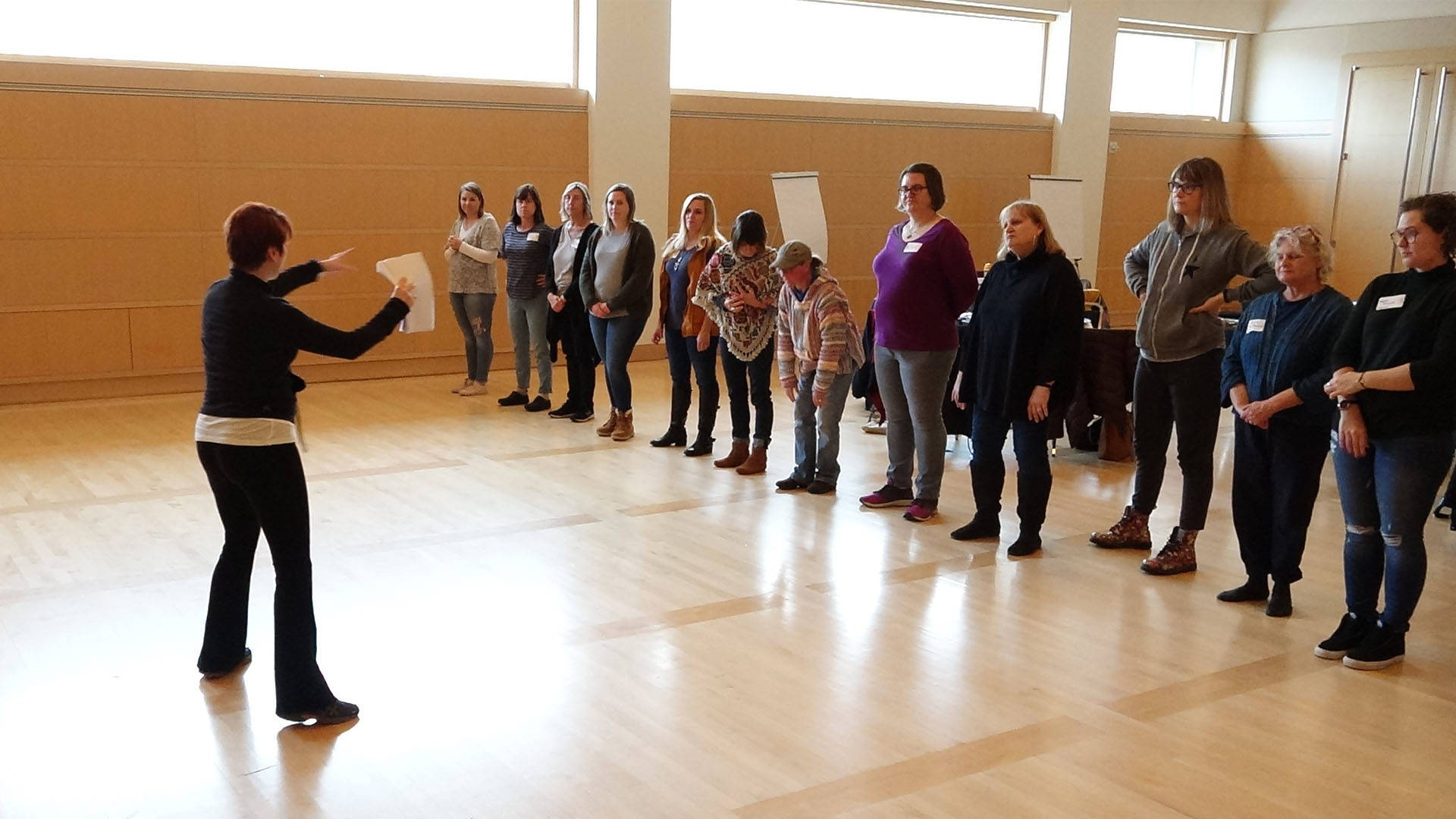 A group of adults standing in a line, while a woman faces them holding a piece of paper and directing them.