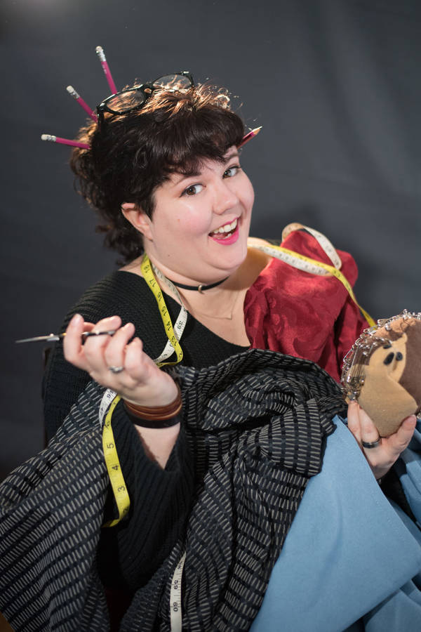 A young white woman with short brown hair covered in various fabrics, tape measures, and sewing tools. 