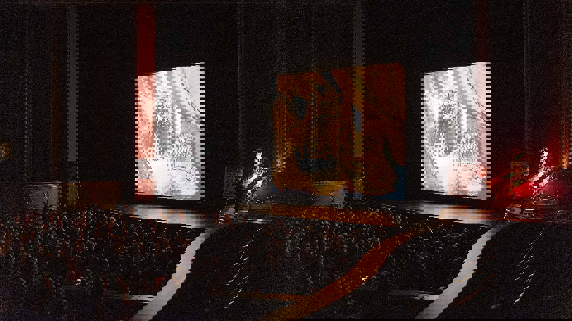 An audience in the orchestra section of Capitol Theater seated and watching a screening of a silent film. An organist plays the red and gold Grand Barton Organ right of the stage.