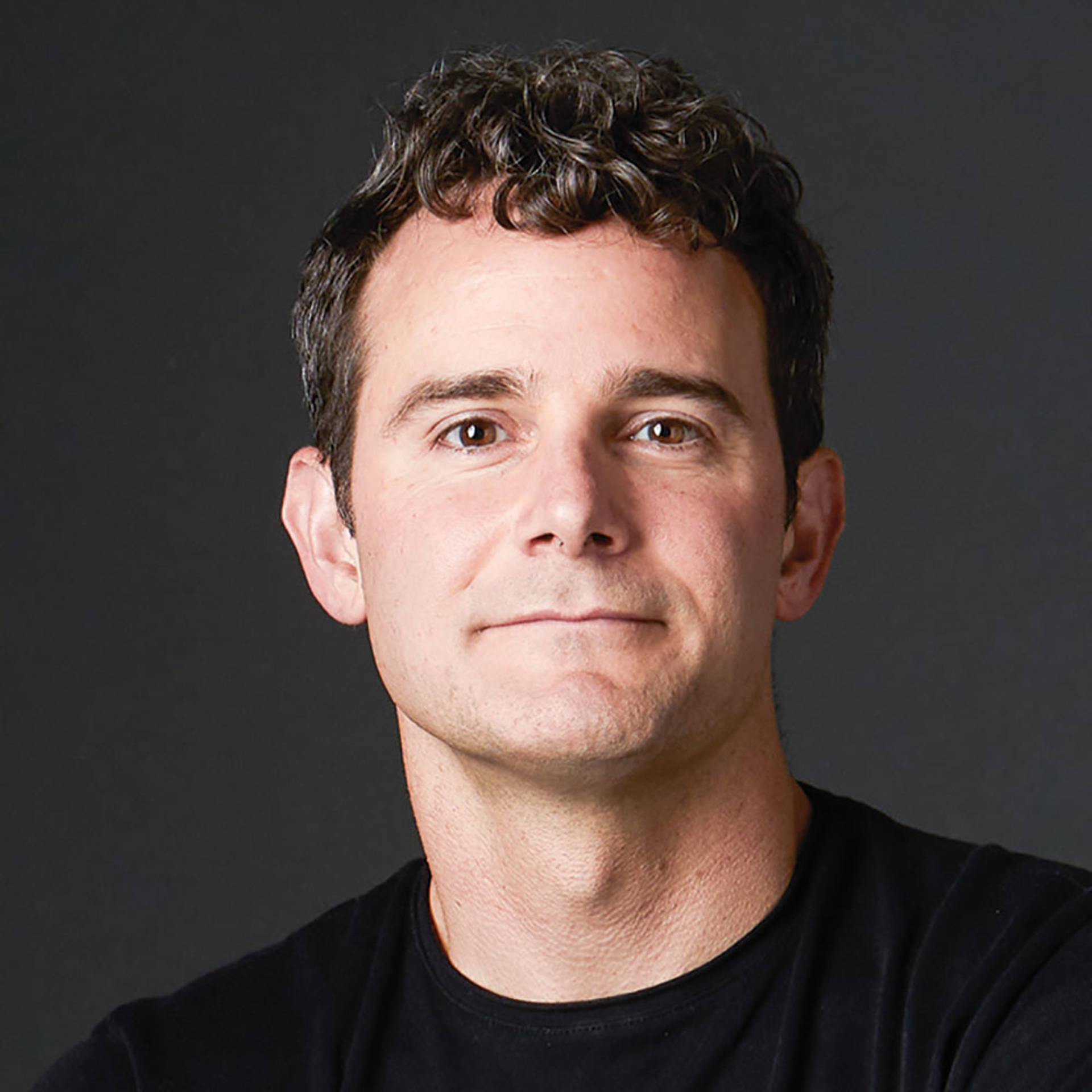 Headshot of a white man with short curly dark hair with brown eyes and a black t shirt.