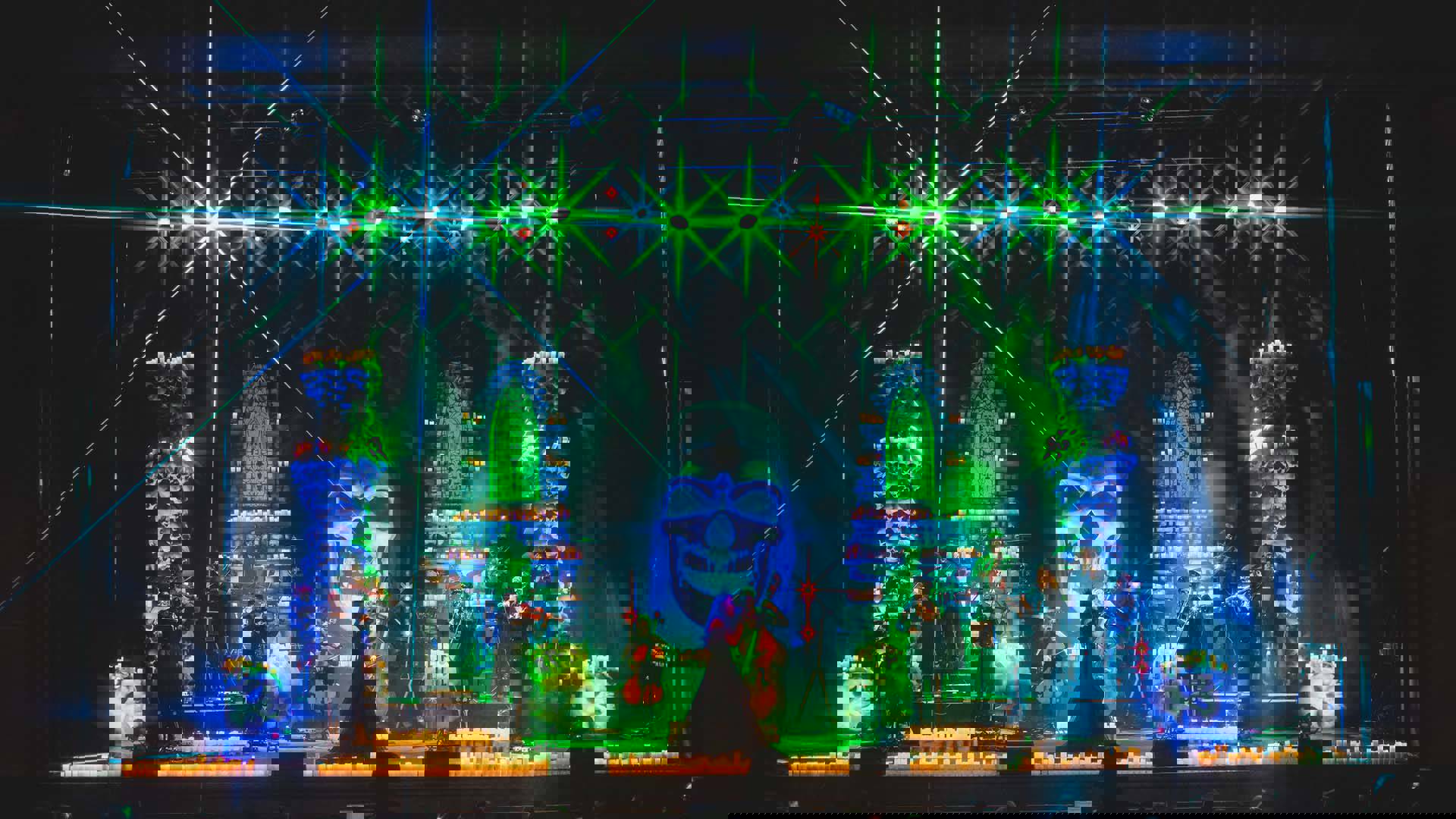 Stage with eleven musical performers, green and blue light beams, and a large skull in background.