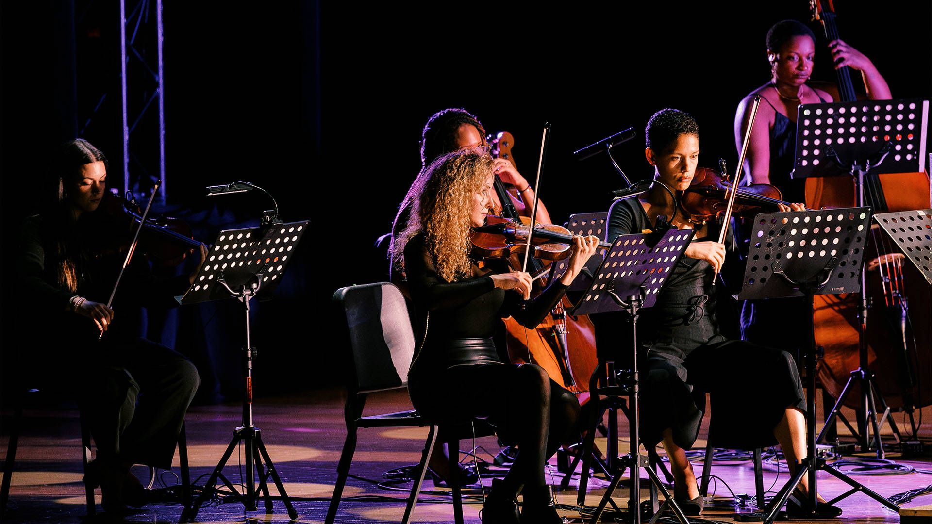 A woman with light complexion, long curly blond hair and a dark outfit playing violin with other string players behind her.